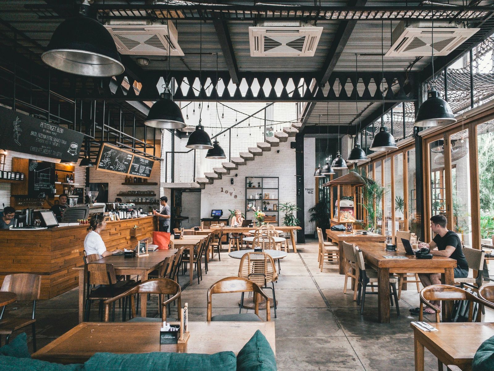 A bright bistro dining room with hanging lamps, wooden tables and a staircase at the back
