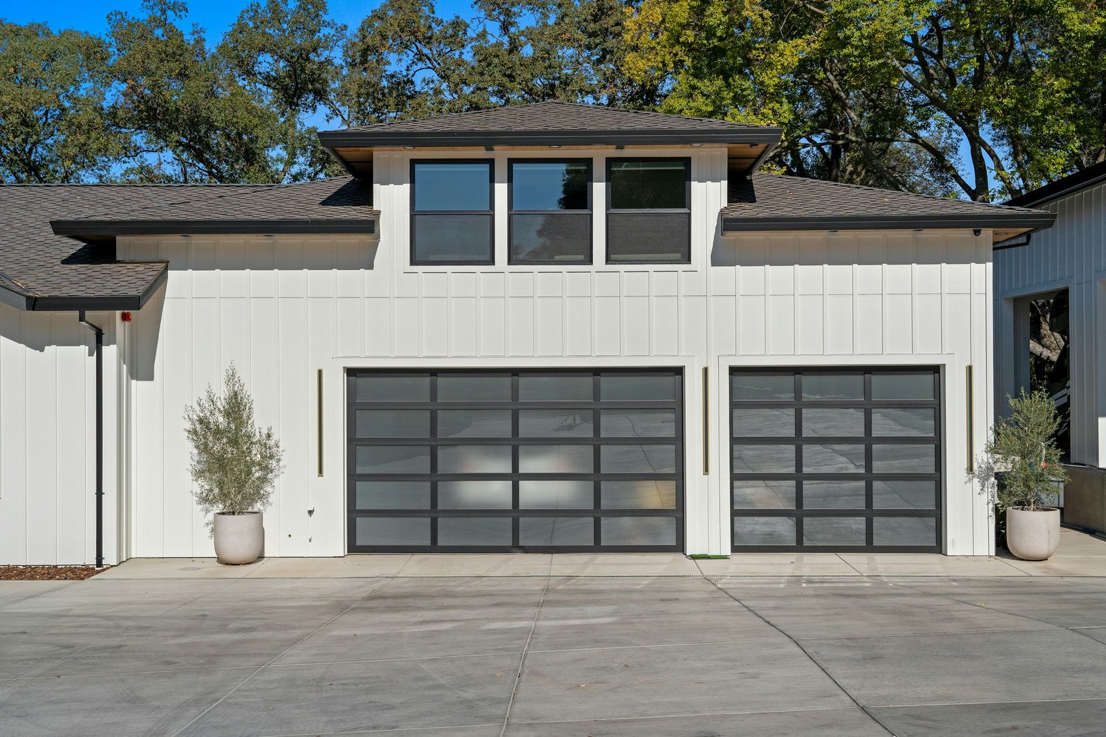 Two dark glass-panel garage doors on a white house, with potted olive trees