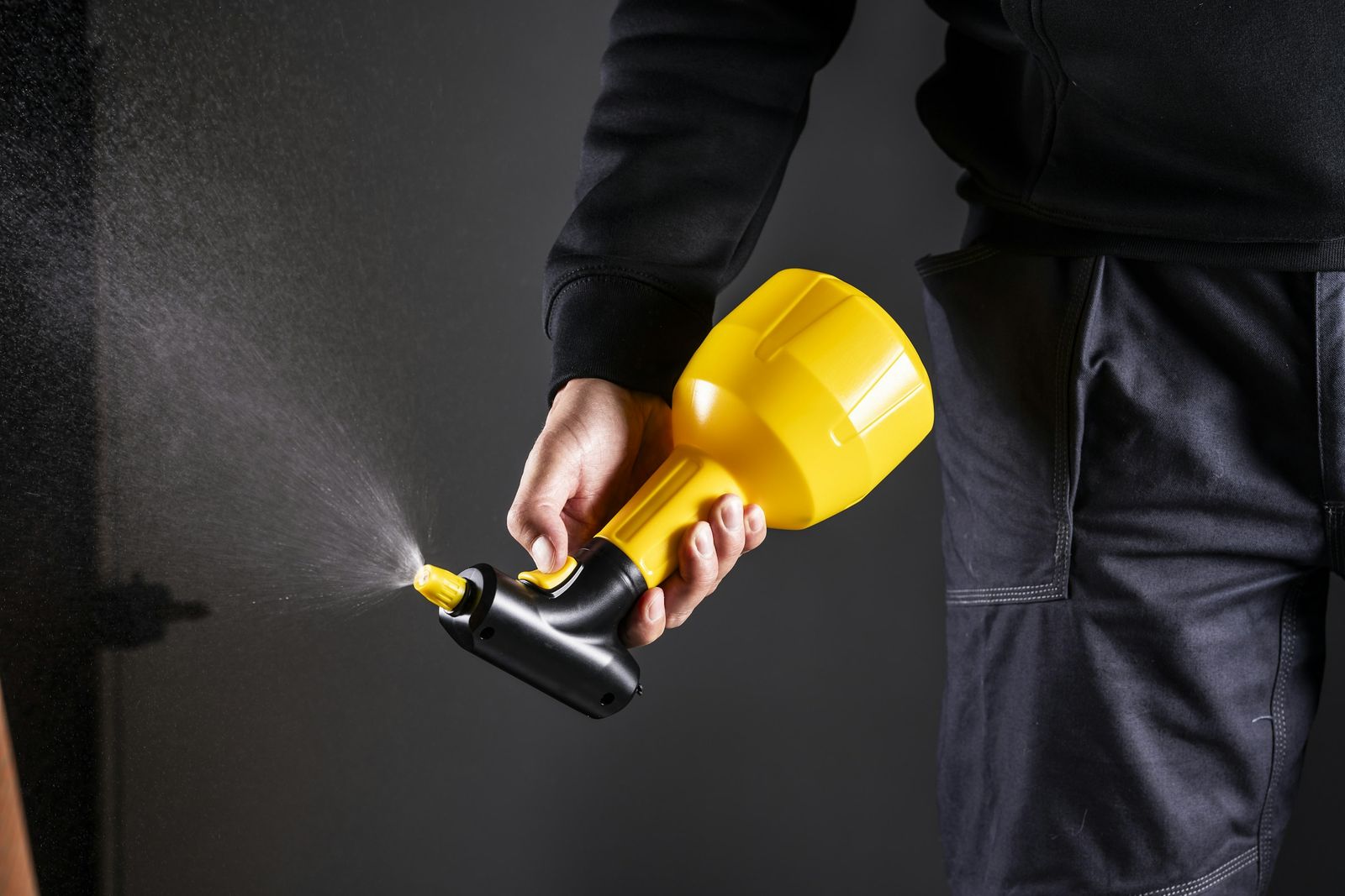 A hand holding a yellow pressure sprayer, misting against a dark background