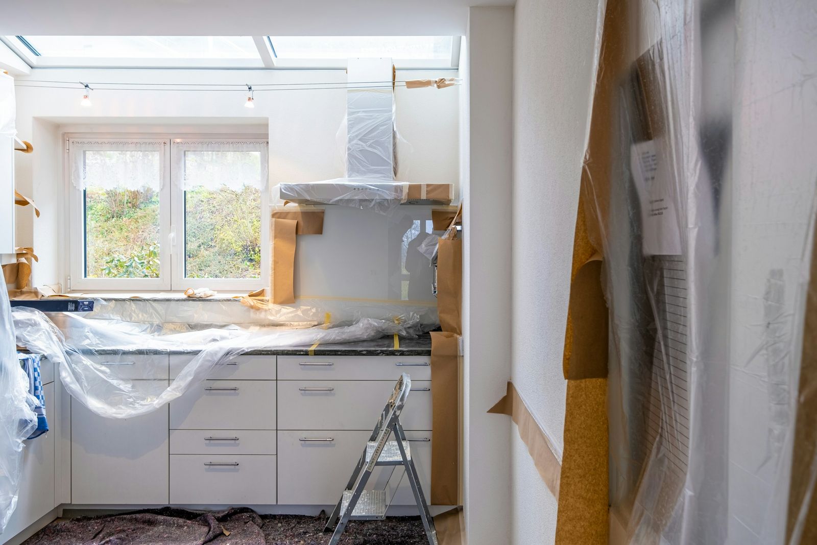 A kitchen under renovation, cabinets and counters wrapped in plastic, a step ladder by the window