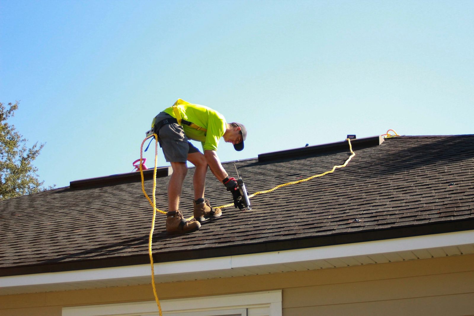 A roofer in a safety harness working on a shingled roof under a blue sky