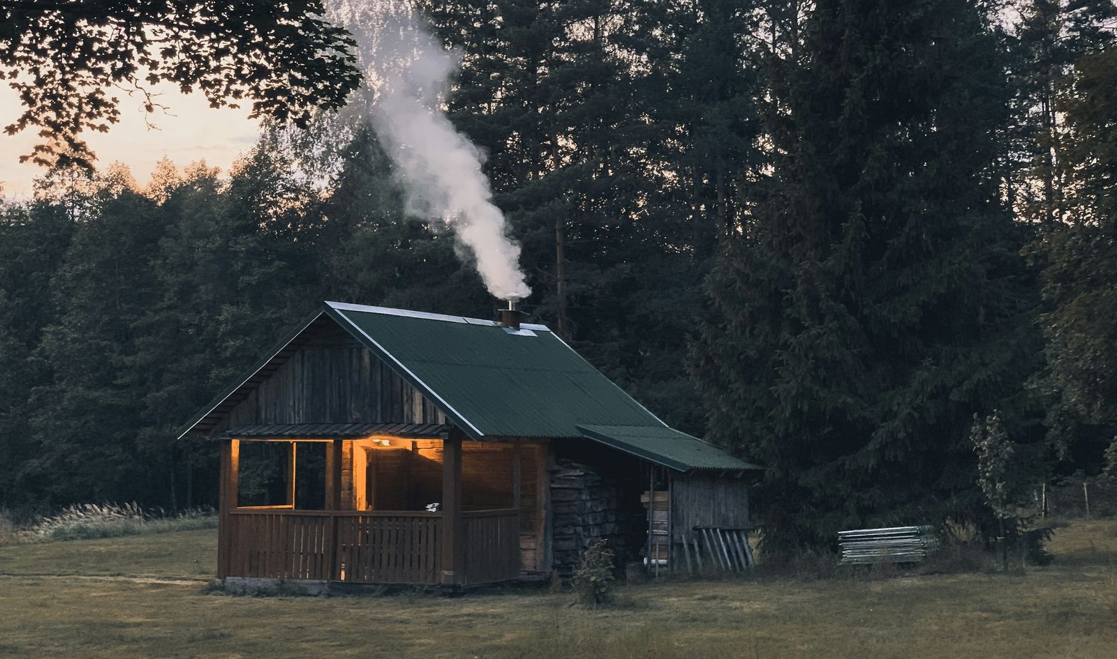A wooden cabin with a green roof at the edge of a conifer forest, smoke rising from the chimney and the porch lit