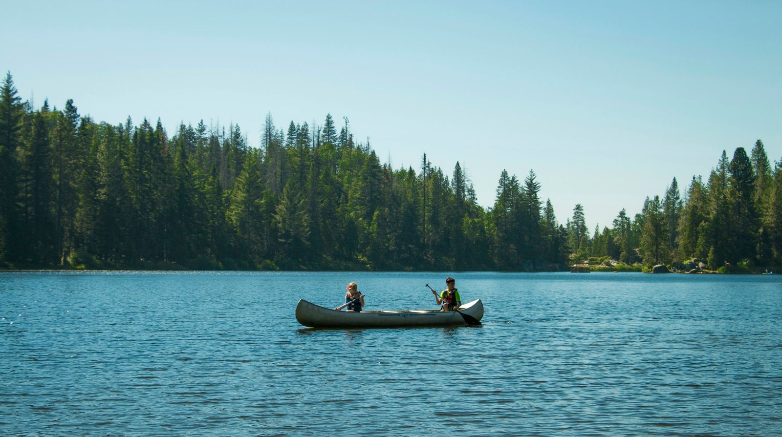 Two paddlers in life jackets in a canoe on a blue lake ringed by conifers
