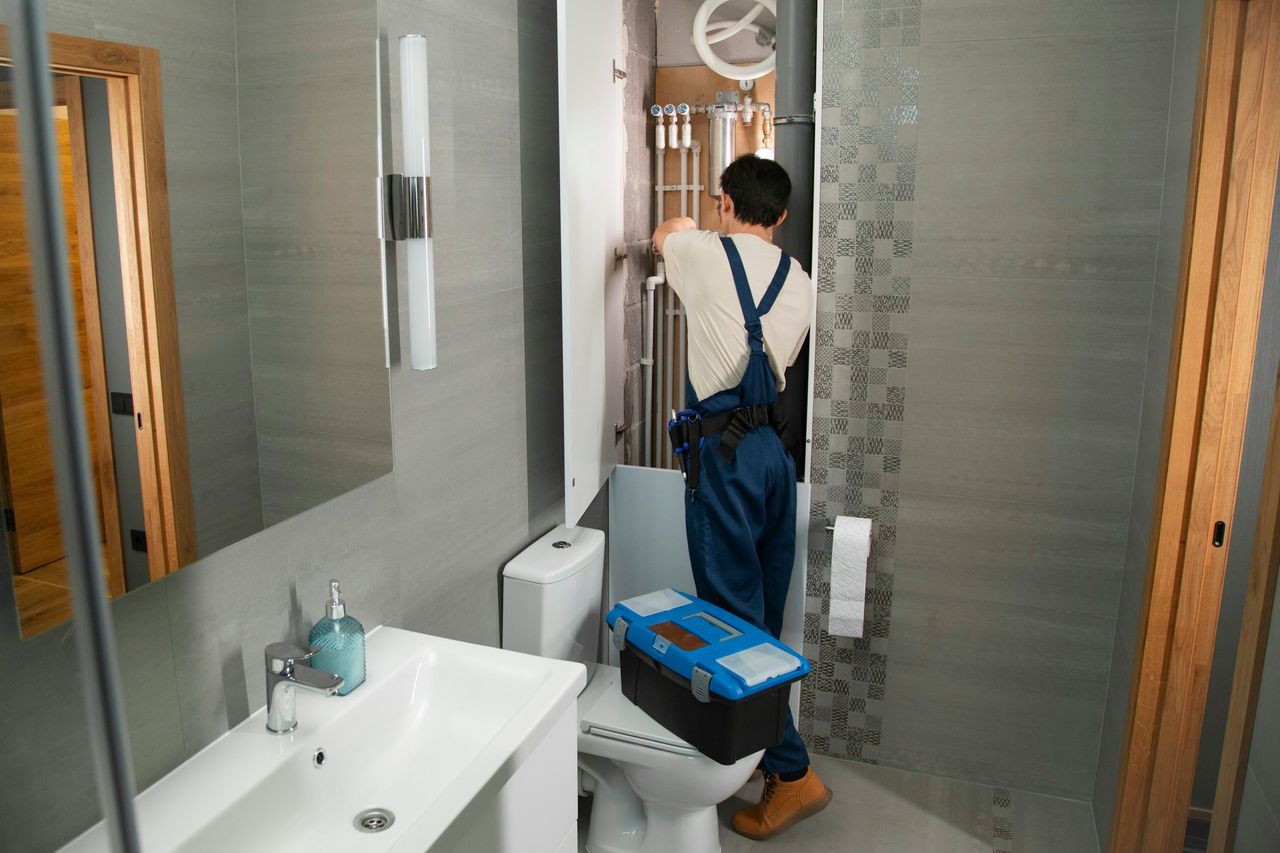 A plumber in overalls working on the pipes behind a bathroom wall panel