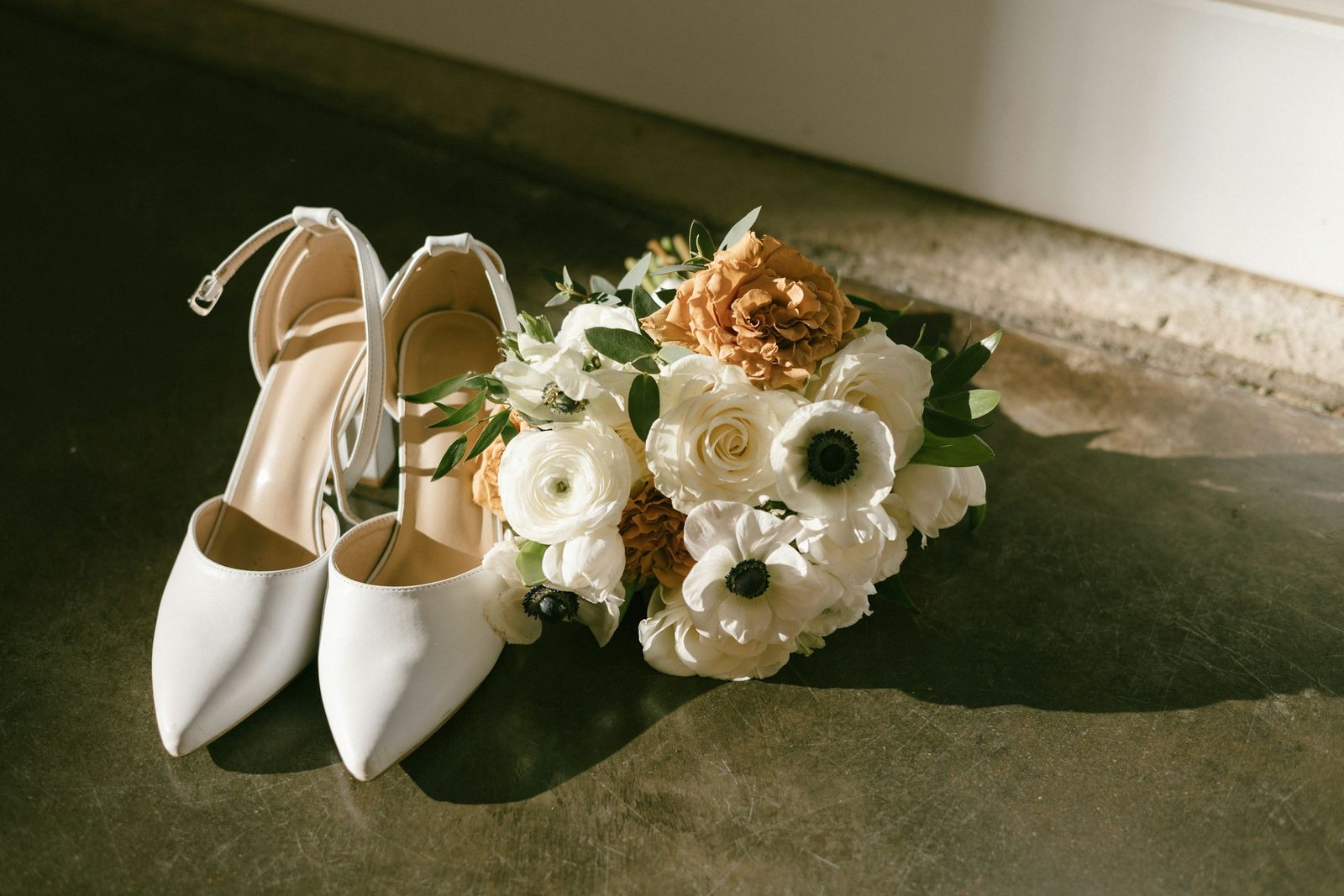 White wedding shoes beside a bouquet of white anemones and roses, in low sunlight